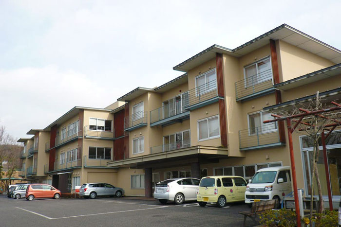 クリーム色と赤褐色の3階建て介護施設。駐車場に複数台の車が停まっている。