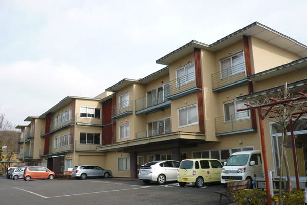 介護施設の建物外観。ベージュと赤茶色の外壁、3階建て、駐車場完備。
